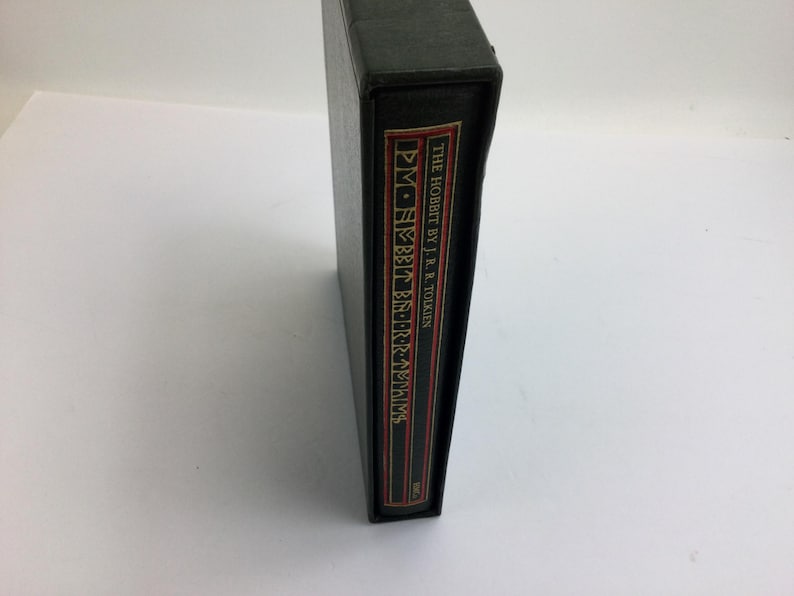 May include: A black book with the title "THE HOBBIT BY J.R.R. TOLKIEN" in white text on a red and gold label. The book is in a black case.
