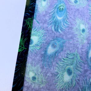 May include: Close-up of fabric with a peacock feather pattern in shades of blue and green on a lavender background. The edge of the fabric has a dark blue border with green accents.