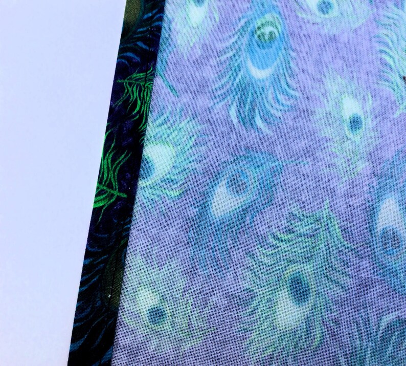 May include: Close-up of fabric with a peacock feather pattern in shades of blue and green on a lavender background. The edge of the fabric has a dark blue border with green accents.