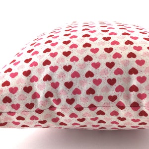 May include: A square throw pillow featuring a repeating pattern of small hearts in shades of red and pink on a white background. The fabric has a subtle speckled texture throughout, and the pillow is shown from a side angle against a white background.