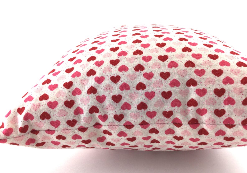 May include: A square throw pillow featuring a repeating pattern of small hearts in shades of red and pink on a white background. The fabric has a subtle speckled texture throughout, and the pillow is shown from a side angle against a white background.