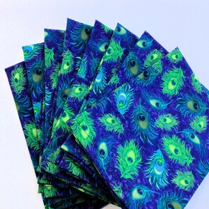 May include: A stack of square fabric napkins with a vibrant peacock feather pattern. The design features bright green and teal feathers on a deep blue background. The napkins are neatly arranged, showcasing the repeating motif.