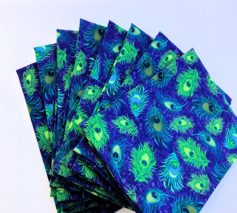 May include: A stack of square fabric napkins with a vibrant peacock feather pattern. The design features bright green and teal feathers on a deep blue background. The napkins are neatly arranged, showcasing the repeating motif.