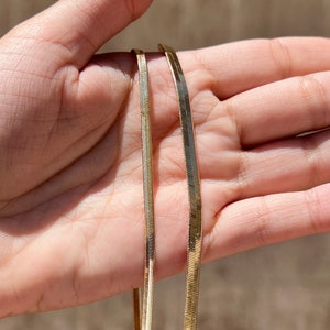 May include: Two gold chain necklaces, one thicker than the other, are displayed on a hand. The chains are both flat and have a shiny finish.