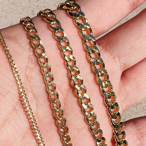May include: Four gold chain necklaces of varying widths, displayed on a hand.