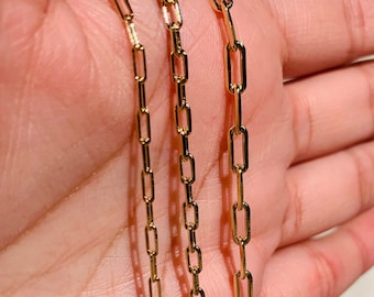 Pode incluir: Três colares de corrente de ouro com um design de corrente de elo quadrado. As correntes estão sobrepostas e são seguradas em uma mão.