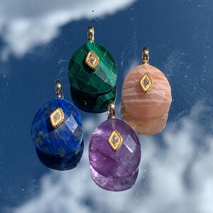 May include: Four gemstone pendants in shades of blue, green, purple, and peach. Each pendant has a gold-colored bail and a small diamond-like accent. The pendants are displayed against a reflective surface with a sky and cloud background.