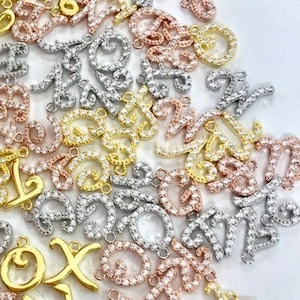 May include: A large assortment of cursive letter charms in gold, silver, and rose gold. Each charm is adorned with small clear cubic zirconia stones.