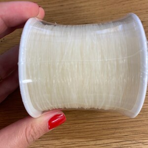 Puede incluir: Una bobina de hilo transparente y fino, enrollado firmemente y encerrado en un soporte de pl&aacute;stico blanco. El hilo parece ser de nailon o un material similar, ideal para coser o proyectos de manualidades.