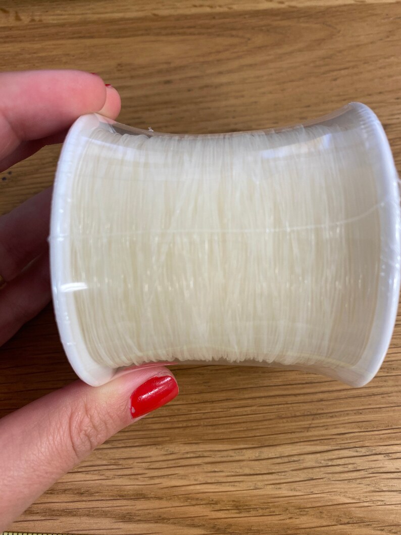 Puede incluir: Una bobina de hilo transparente y fino, enrollado firmemente y encerrado en un soporte de pl&aacute;stico blanco. El hilo parece ser de nailon o un material similar, ideal para coser o proyectos de manualidades.
