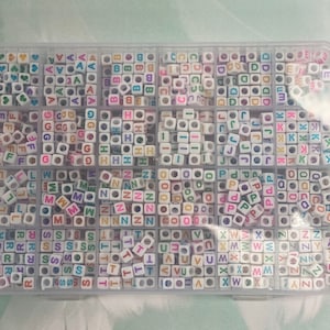 May include: A clear plastic storage box filled with small white letter beads. Each compartment contains a different letter of the alphabet, with the letters A through Z represented. The beads are white with colorful letters.