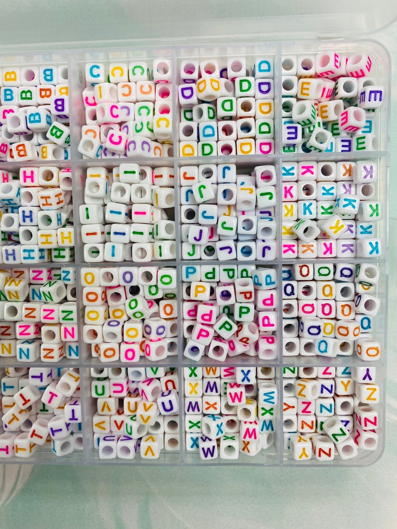 May include: A clear plastic storage box filled with white letter beads. Each compartment contains a different letter of the alphabet, with each letter represented in a variety of bright colors.
