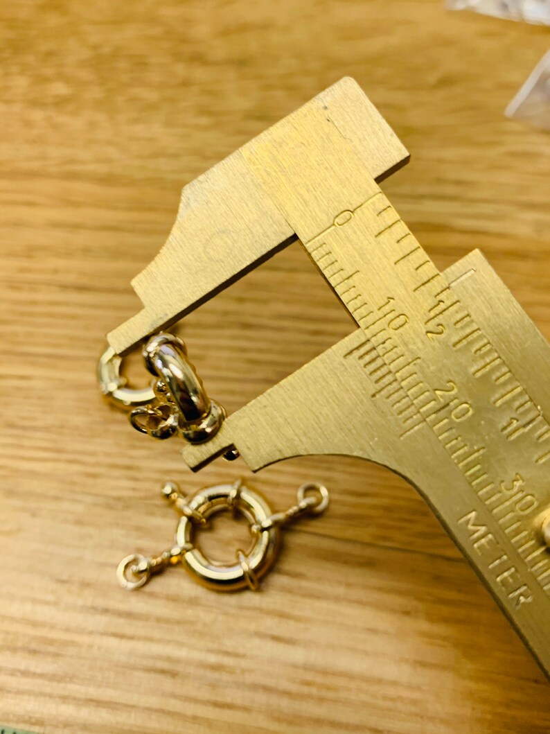 K&ouml;nnte beinhalten: Nahaufnahme von goldfarbenen Schmuckverschl&uuml;ssen und einem Messschieber aus Messing. Die Verschl&uuml;sse umfassen einen runden Federringverschluss und einen kreisf&ouml;rmigen Verschluss mit drei kleinen Armen. Der Messschieber misst die Gr&ouml;&szlig;e des Verschlusses.