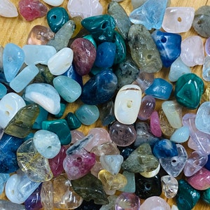 May include: A collection of colorful, natural stone beads with holes drilled through the center. The beads are various shapes and sizes, including round, oval, and irregular shapes. The beads are a mix of colors, including blue, green, pink, purple, and brown.