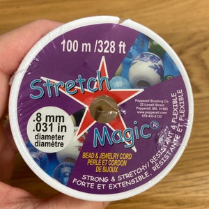 Puede incluir: Un carrete de cord&oacute;n Stretch Magic para cuentas y joyer&iacute;a. La etiqueta es morada y blanca, con el texto "Stretch Magic" en una estrella roja. El cord&oacute;n tiene 0,8 mm de di&aacute;metro y 100 m de largo.