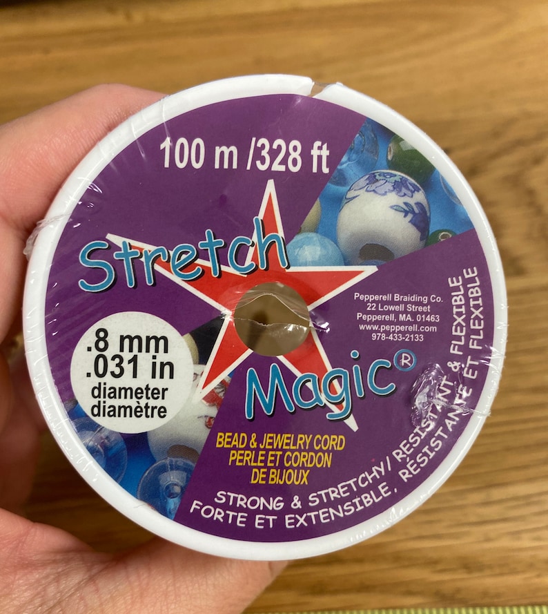 Puede incluir: Un carrete de cord&oacute;n Stretch Magic para cuentas y joyer&iacute;a. La etiqueta es morada y blanca, con el texto "Stretch Magic" en una estrella roja. El cord&oacute;n tiene 0,8 mm de di&aacute;metro y 100 m de largo.