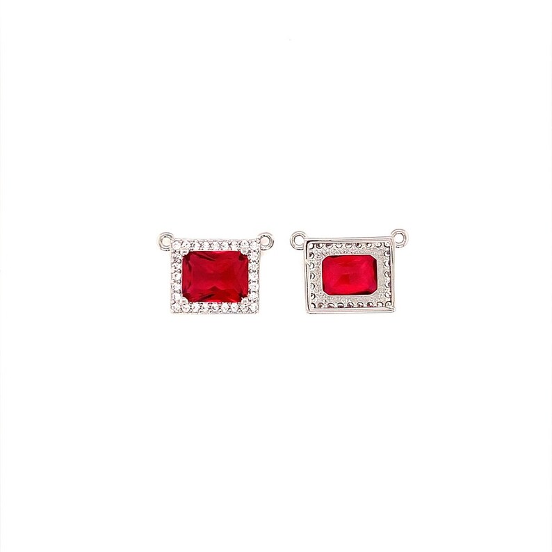 May include: Two silver-toned rectangular pendants with a vibrant red gemstone center. One pendant is surrounded by a halo of small, clear crystals. The other pendant has a decorative border. Both have small loops for attaching to a chain.