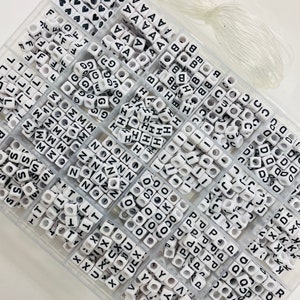 May include: A clear plastic storage box filled with white letter beads. The beads are organized by letter in individual compartments. The box also includes a spool of clear elastic string.