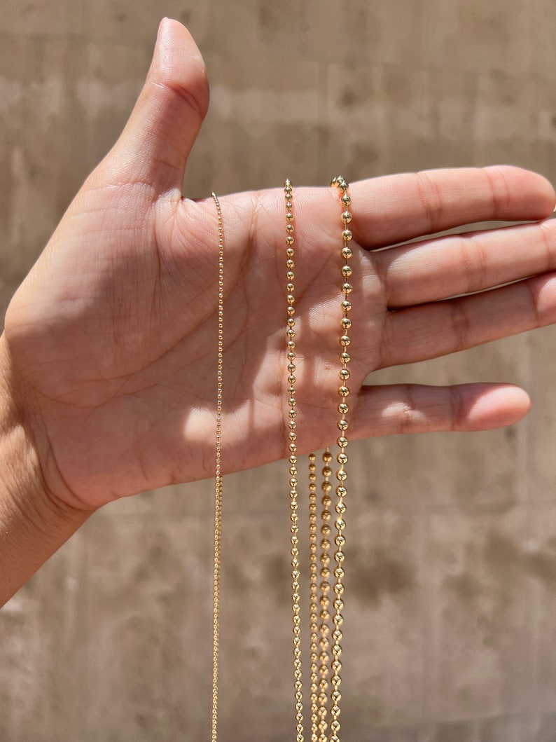 May include: Three gold chains with ball links of different sizes. The chains are laid out on a hand, with the smallest chain on the left and the largest chain on the right.