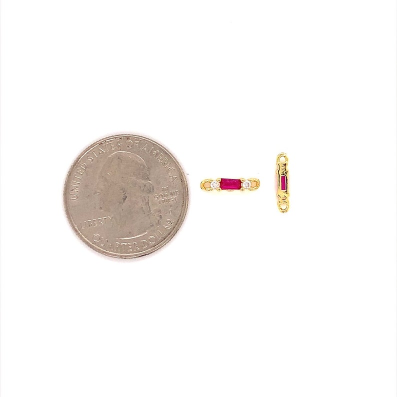 May include: Two small gold-colored jewelry pieces with red and clear stones. One piece is horizontal, the other vertical. A U.S. quarter coin is next to the jewelry for size comparison. The jewelry has a rectangular shape with a red stone in the center and clear stones on each side.