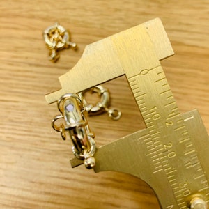 K&ouml;nnte beinhalten: Nahaufnahme von goldfarbenen Schmuckverschl&uuml;ssen und einem Messschieber aus Messing auf einer Holzoberfl&auml;che. Die Verschl&uuml;sse haben ein dekoratives Design und werden f&uuml;r die Schmuckherstellung verwendet. Der Messschieber dient zum Messen der Verschl&uuml;sse.