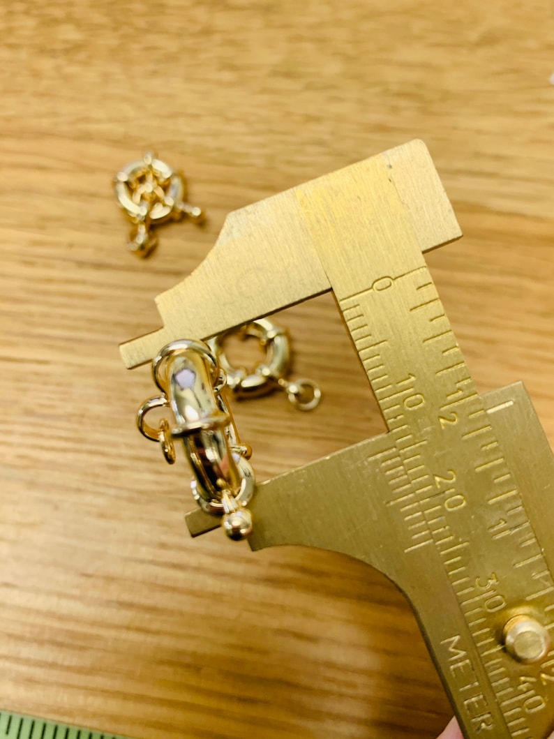 K&ouml;nnte beinhalten: Nahaufnahme von goldfarbenen Schmuckverschl&uuml;ssen und einem Messschieber aus Messing auf einer Holzoberfl&auml;che. Die Verschl&uuml;sse haben ein dekoratives Design und werden f&uuml;r die Schmuckherstellung verwendet. Der Messschieber dient zum Messen der Verschl&uuml;sse.
