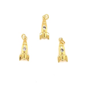 Puede incluir: Tres colgantes dorados con forma de cohete, cada uno con una pequeña gema transparente en el centro. Los colgantes tienen un lazo en la parte superior para sujetarlos a un collar o pulsera. Los cohetes tienen un diseño clásico con aletas en la base.