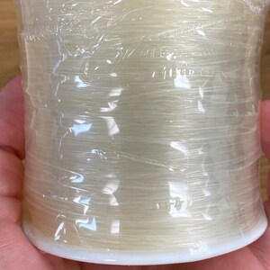 Puede incluir: Un carrete de hilo de pescar monofilamento transparente y fino, envuelto en pl&aacute;stico transparente. El hilo es de un color p&aacute;lido y transl&uacute;cido, enrollado alrededor de una bobina de pl&aacute;stico blanco. El envoltorio de pl&aacute;stico est&aacute; arrugado.