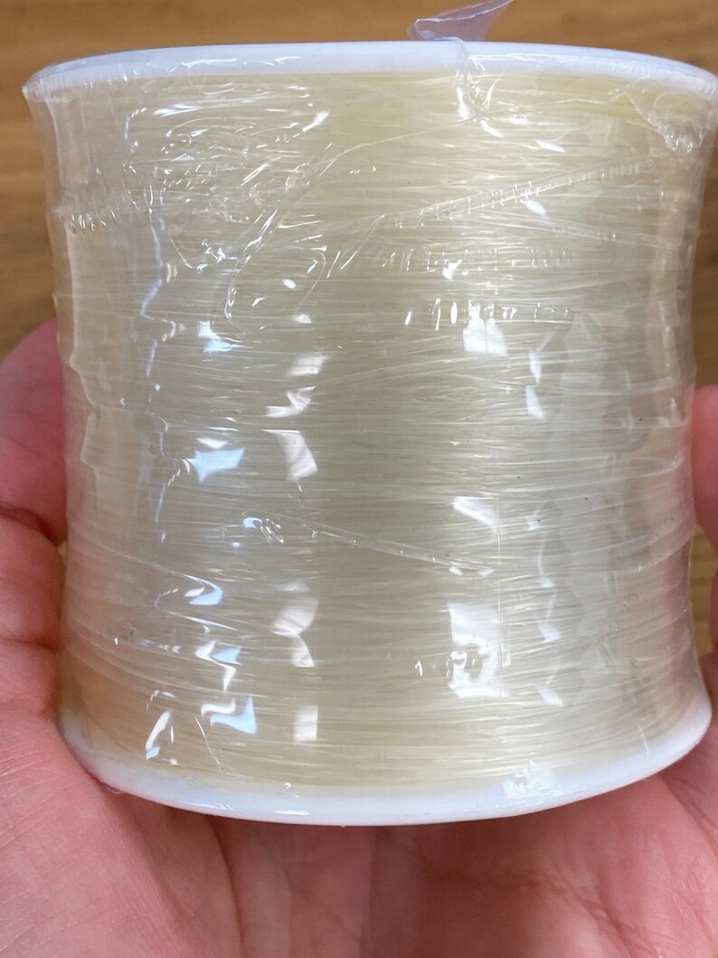 Puede incluir: Un carrete de hilo de pescar monofilamento transparente y fino, envuelto en pl&aacute;stico transparente. El hilo es de un color p&aacute;lido y transl&uacute;cido, enrollado alrededor de una bobina de pl&aacute;stico blanco. El envoltorio de pl&aacute;stico est&aacute; arrugado.