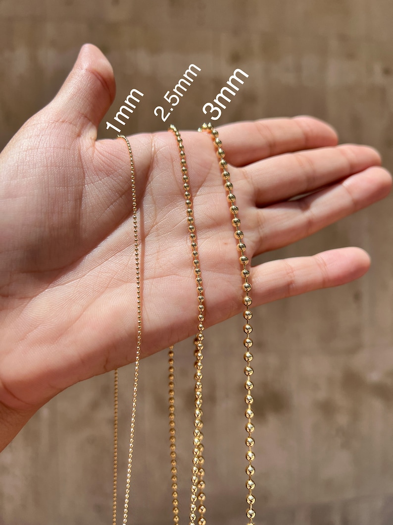 May include: Three gold chains of different sizes, 1mm, 2.5mm, and 3mm, are shown on a hand. The chains are made of small, round beads.