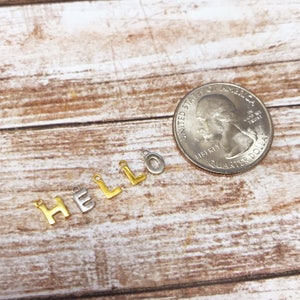 May include: Gold and silver letter charms spelling out the word "HELLO" on a wooden surface. The charms are about the size of a US quarter.