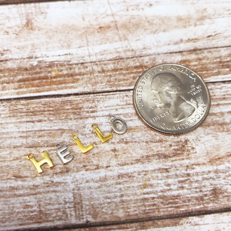 May include: Gold and silver letter charms spelling out the word "HELLO" on a wooden surface. The charms are about the size of a US quarter.