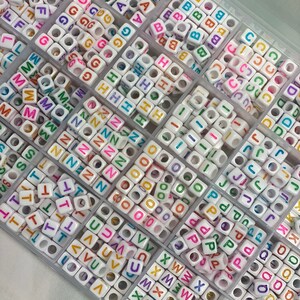 May include: A clear plastic storage box filled with white letter beads. The beads are arranged in rows by letter and have colorful letters on them.