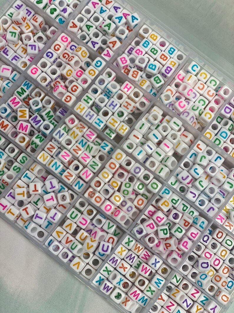 May include: A clear plastic storage box filled with white letter beads. The beads are arranged in rows by letter and have colorful letters on them.