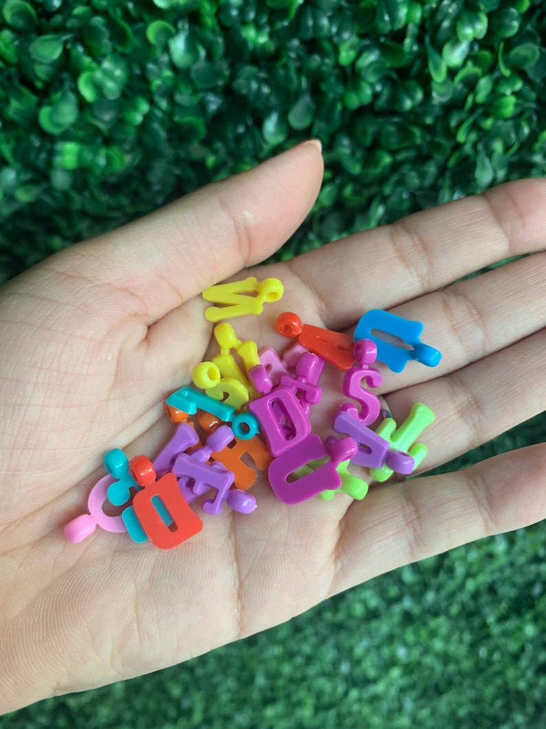 May include: A handful of colorful plastic letter beads in various colors, including pink, purple, yellow, orange, blue, and green. The beads are in the shape of uppercase letters.