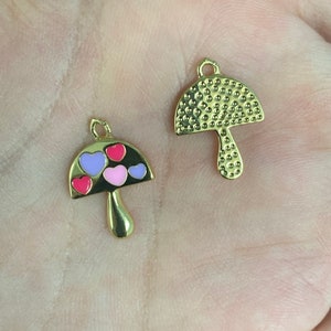 May include: Two gold-colored mushroom charms. One charm has a textured cap and stem. The other has a black cap with pink, purple, and red heart-shaped accents. Both charms have a small loop at the top for attaching to jewelry.