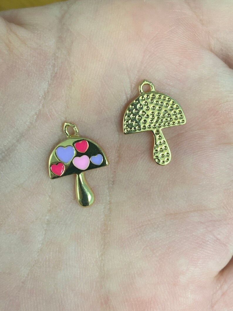 May include: Two gold-colored mushroom charms. One charm has a textured cap and stem. The other has a black cap with pink, purple, and red heart-shaped accents. Both charms have a small loop at the top for attaching to jewelry.
