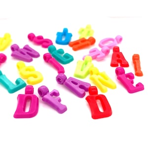 May include: Assortment of colorful plastic letter charms in various colors, including pink, purple, red, orange, yellow, green, blue, and turquoise. The letters are in uppercase and are scattered on a white background.