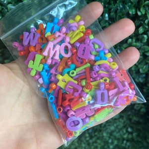 May include: A plastic bag filled with colorful letter beads. The beads are in various colors, including red, yellow, blue, green, and purple. The beads are in the shape of letters of the alphabet.