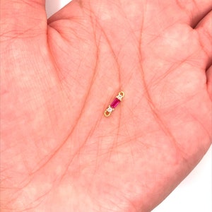 May include: A small, gold-colored jewelry piece with a rectangular, deep pink gemstone in the center, flanked by two smaller, clear gemstones. The piece is held in a person's hand.