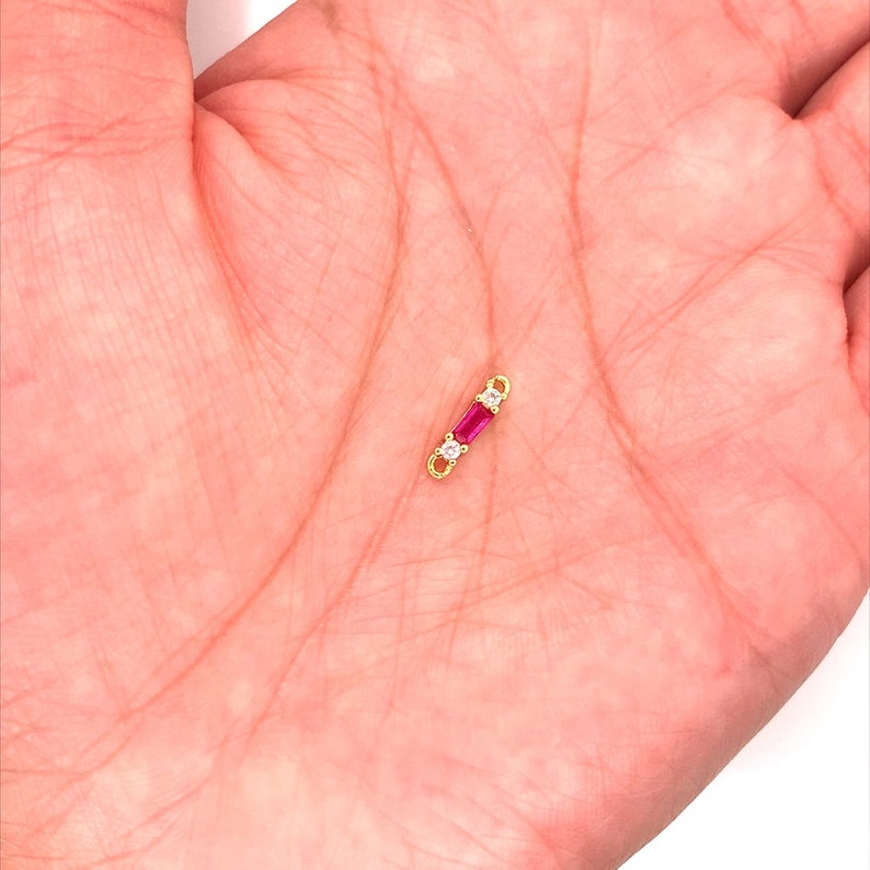 May include: A small, gold-colored jewelry piece with a rectangular, deep pink gemstone in the center, flanked by two smaller, clear gemstones. The piece is held in a person's hand.