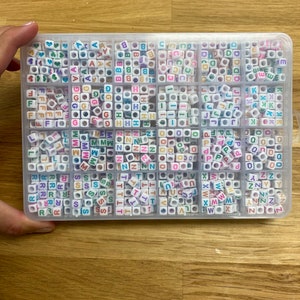 May include: A clear plastic storage box filled with colorful letter beads. The beads are arranged in alphabetical order and are in various colors, including pink, blue, green, yellow, and orange.