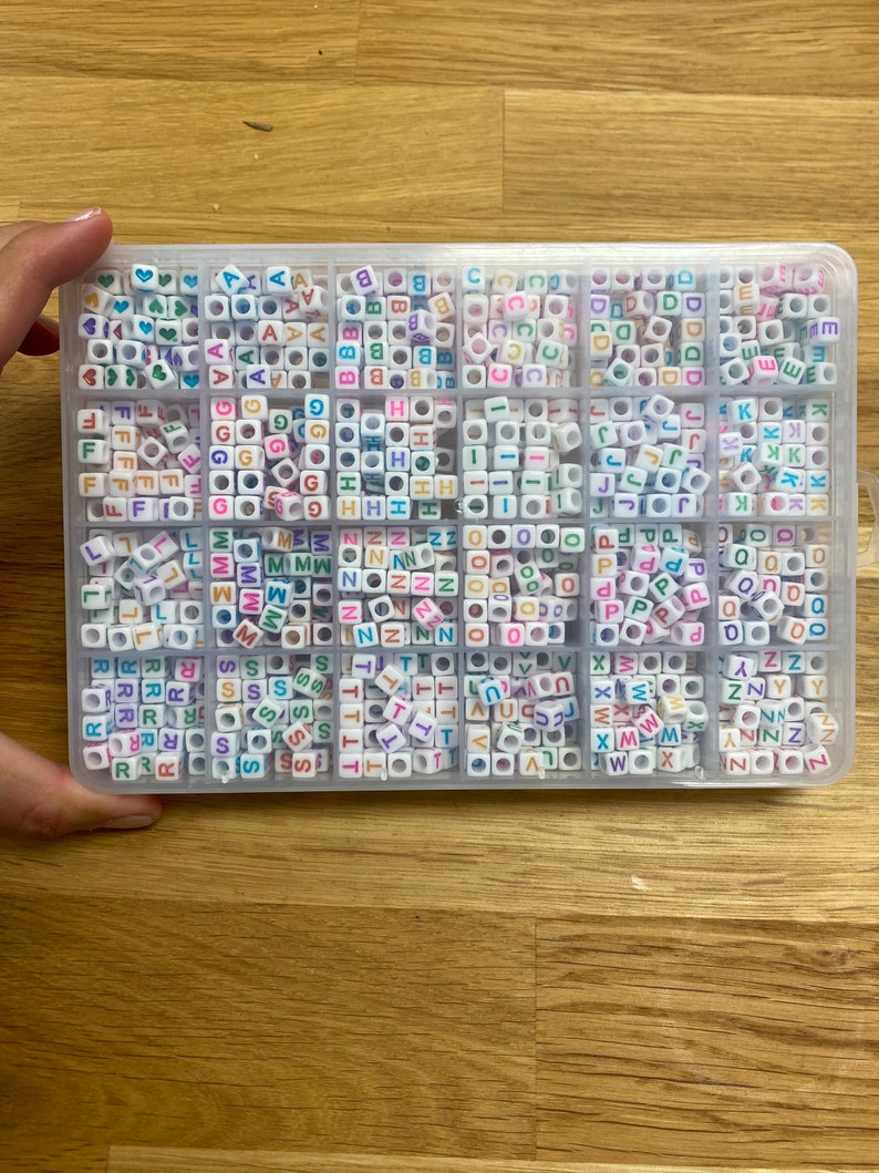 May include: A clear plastic storage box filled with colorful letter beads. The beads are arranged in alphabetical order and are in various colors, including pink, blue, green, yellow, and orange.