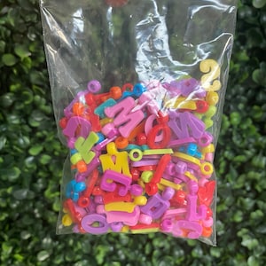 May include: A plastic bag filled with colorful letter and shape beads. The beads are in various colors, including pink, purple, yellow, green, red, and blue.
