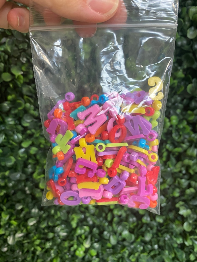 May include: A plastic bag filled with colorful letter and shape beads. The beads are in various colors, including pink, purple, yellow, green, red, and blue.