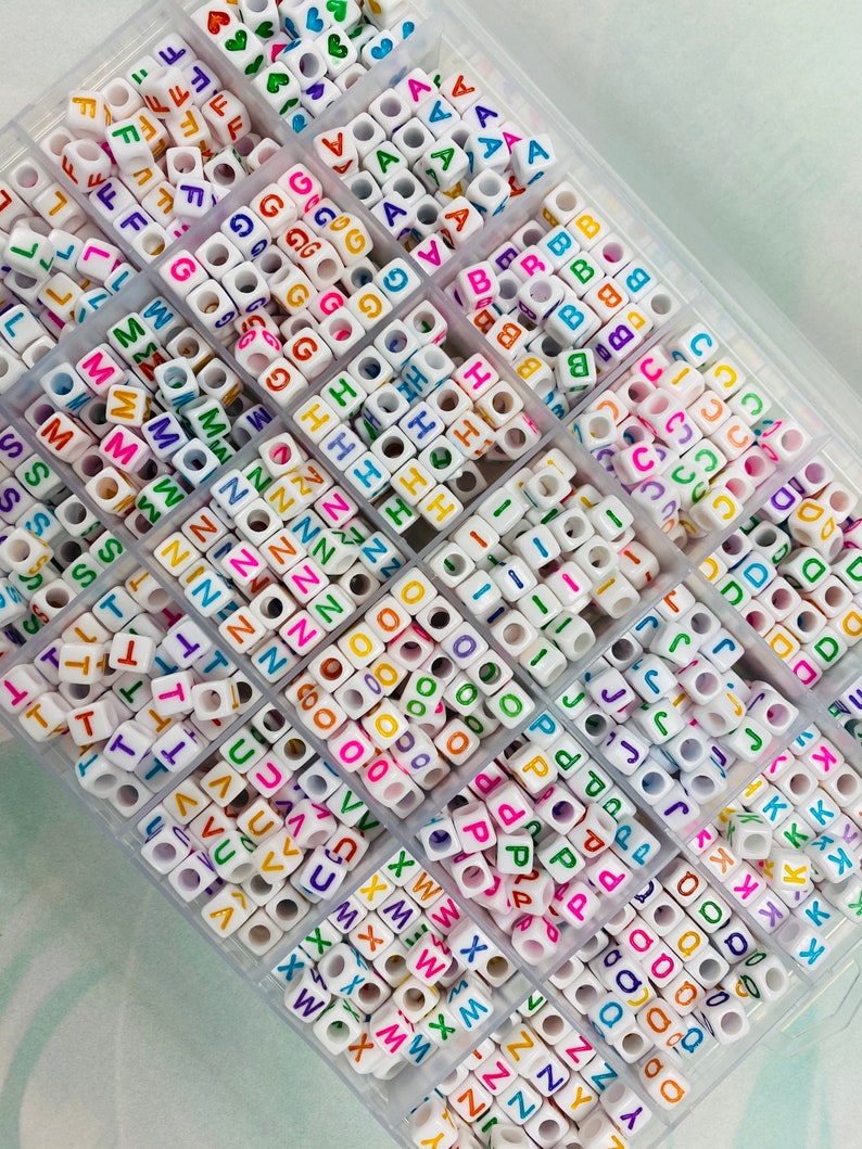 May include: A clear plastic storage box filled with white letter beads. Each compartment contains a different letter of the alphabet, with the letters A through Z represented. The beads are colorful, with each letter printed in a different color.