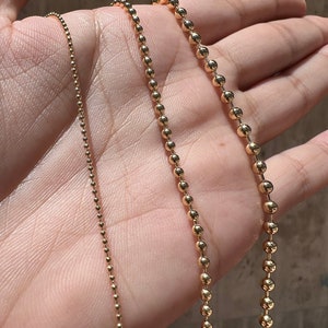 May include: Three gold chains of different thicknesses, all with a ball chain design, are displayed on a hand.