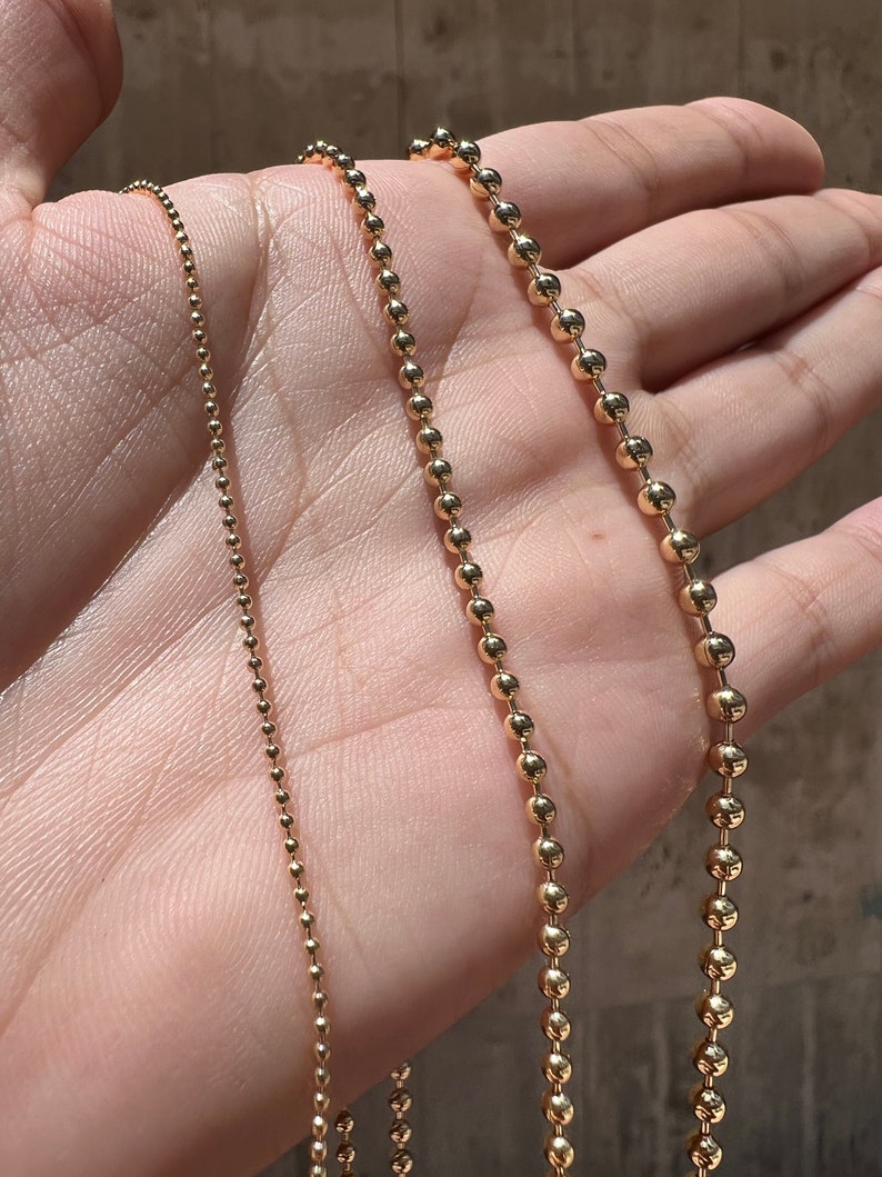 May include: Three gold chains of different thicknesses, all with a ball chain design, are displayed on a hand.