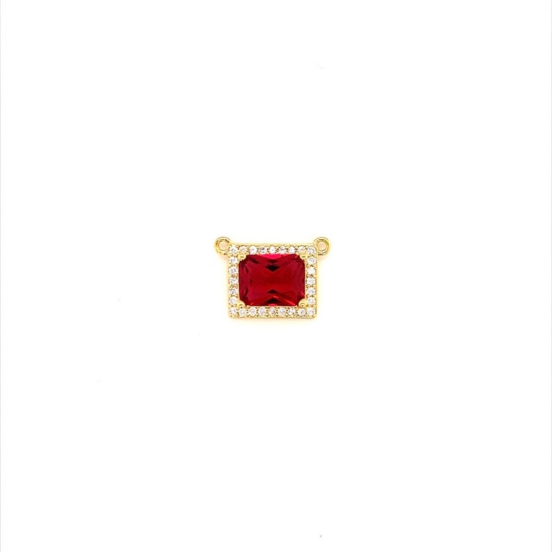 May include: A gold-colored pendant featuring a rectangular, deep red gemstone, surrounded by a halo of smaller, clear stones. The pendant has two small loops at the top for a chain.