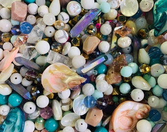 May include: A close-up of a mix of beads in different shapes and colours. The beads include white, turquoise, gold, and iridescent crystals. Some are round, others are teardrop-shaped or faceted. The beads are scattered, creating a textured surface.
